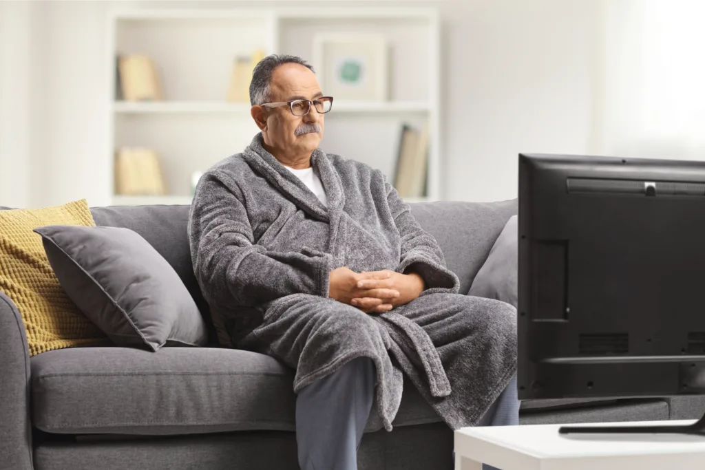 Mature man wearing a bathrobe, sitting on a gray sofa and watchin tv at home