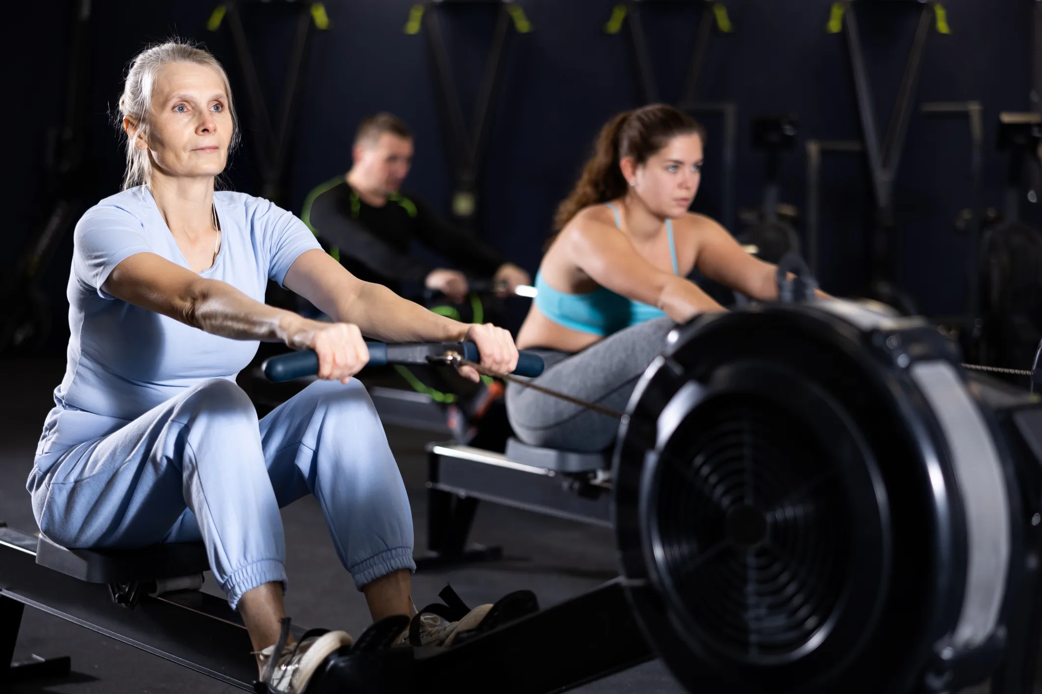 Concentrated sporty senior woman working out on rowing machine during total-body workout in gym. Active lifestyle of older adults concept