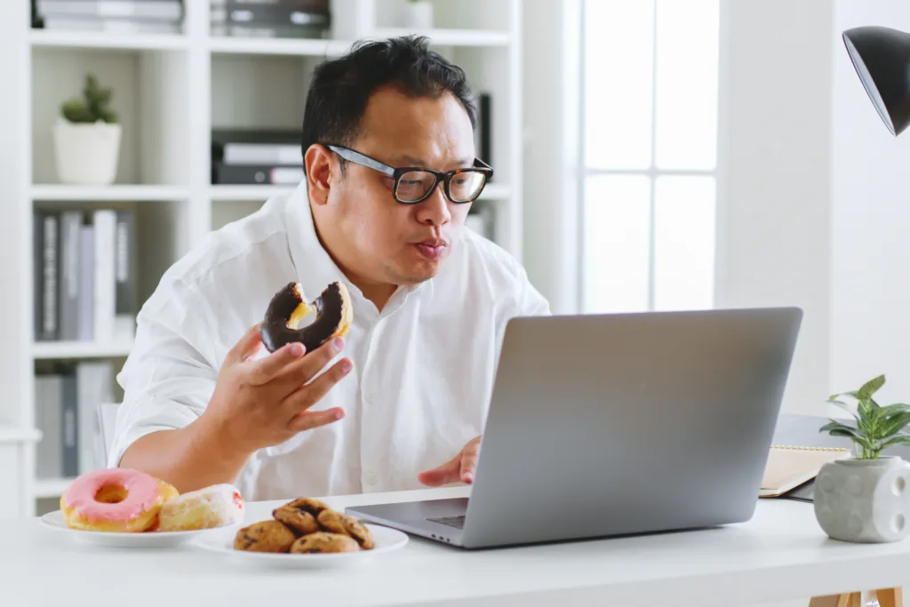 Asian fat man eating donut, sweet, junk food during working with computer laptop, unhealthy eating concept