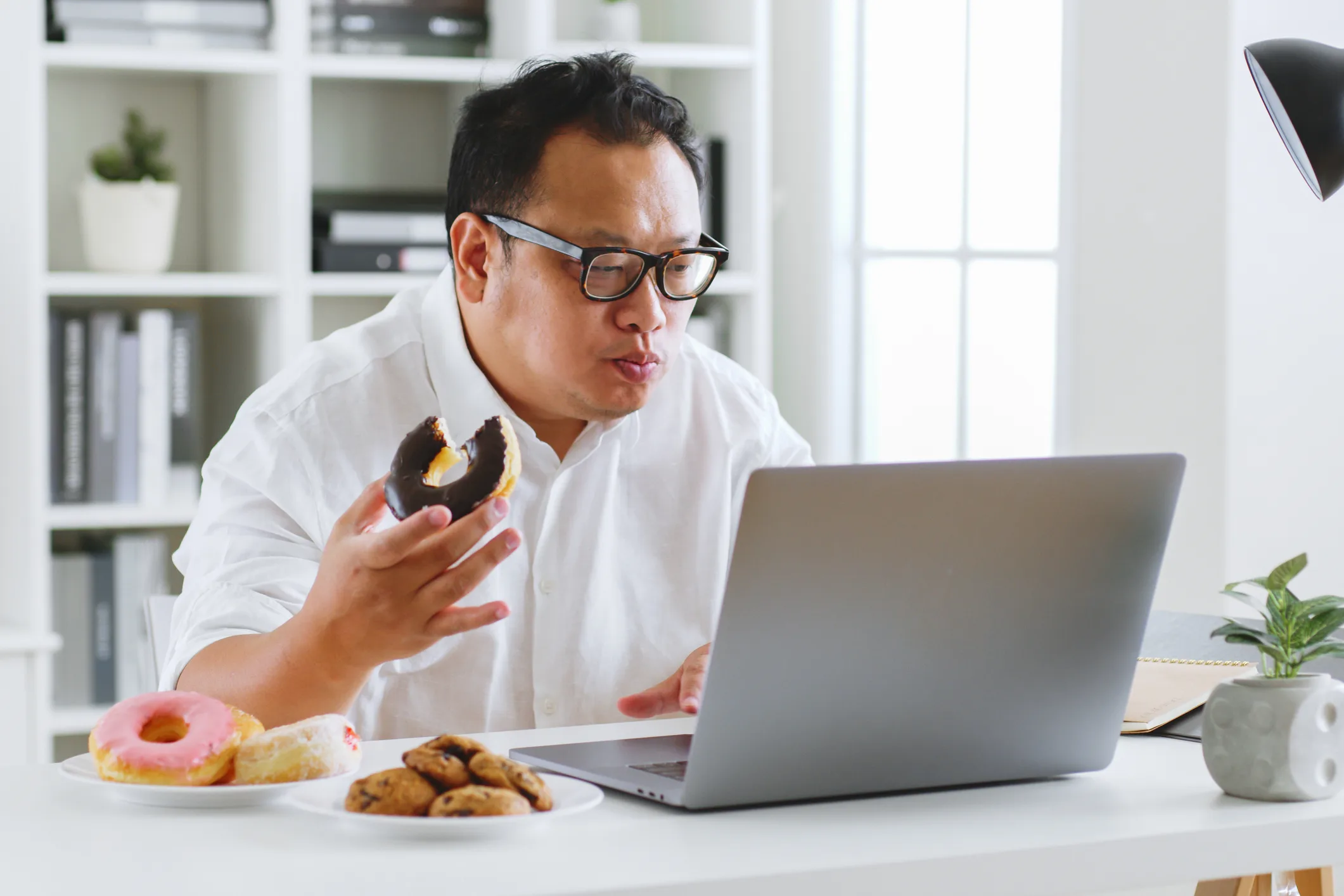 Asian fat man eating donut, sweet, junk food during working with computer laptop, unhealthy eating concept