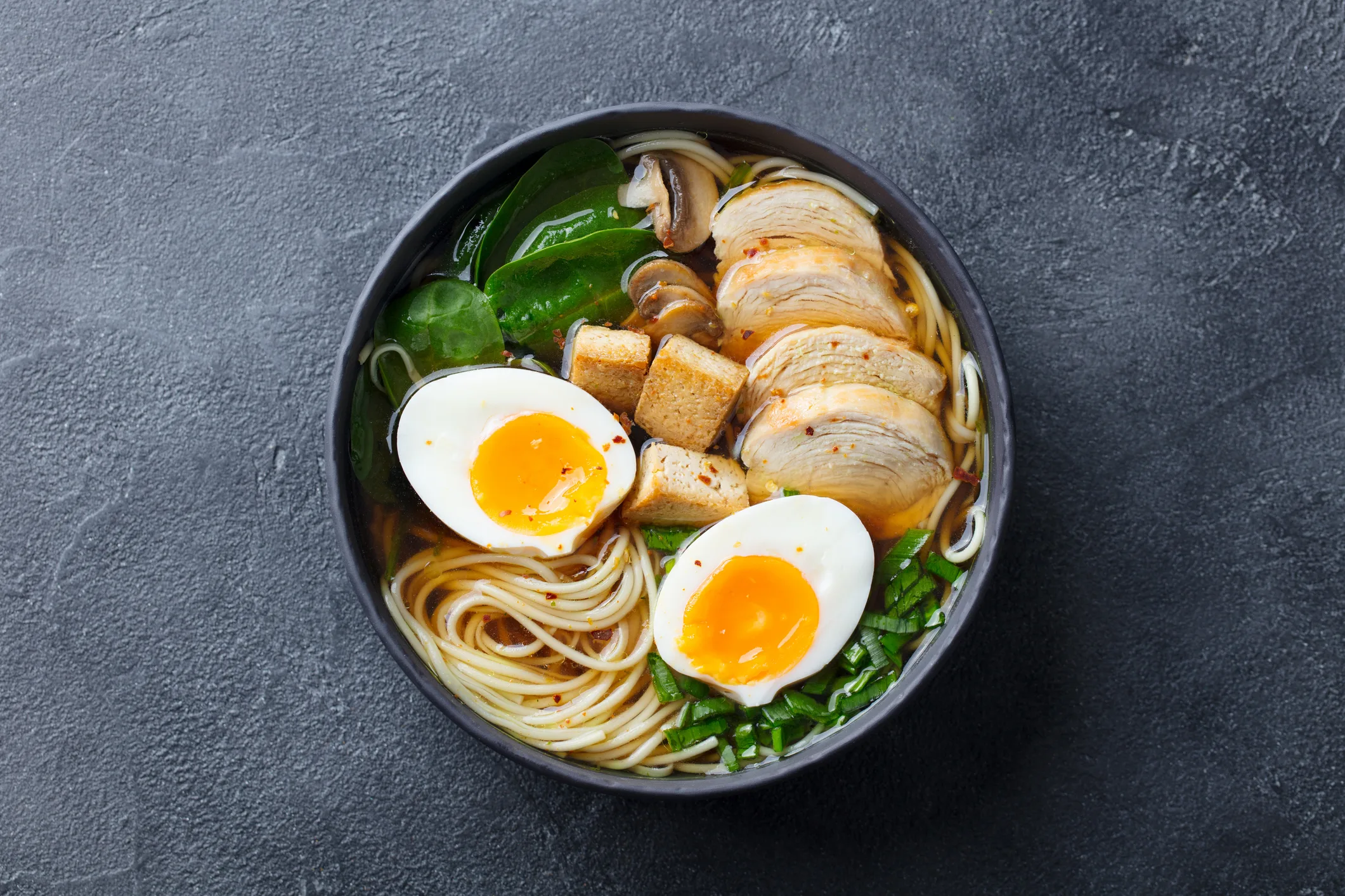 Asian noodle soup, ramen with chicken, tofu, vegetables and egg in black bowl. Top view. Copy space. Slate background.