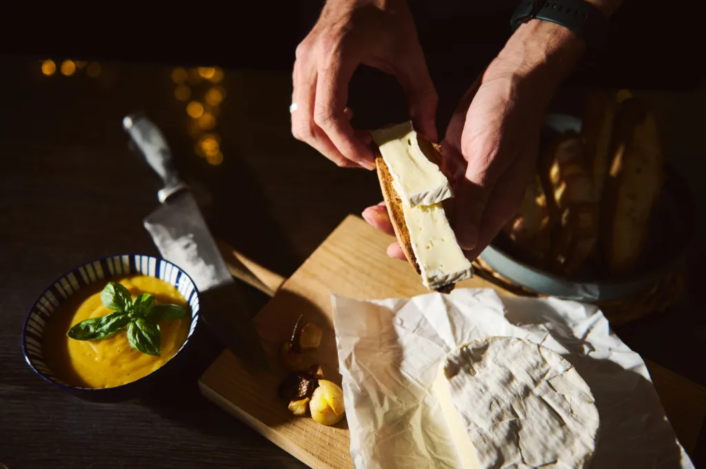 Hands Buttering Bread With Cream Cheese On a Wooden Board Beside Soup and Bread Basket