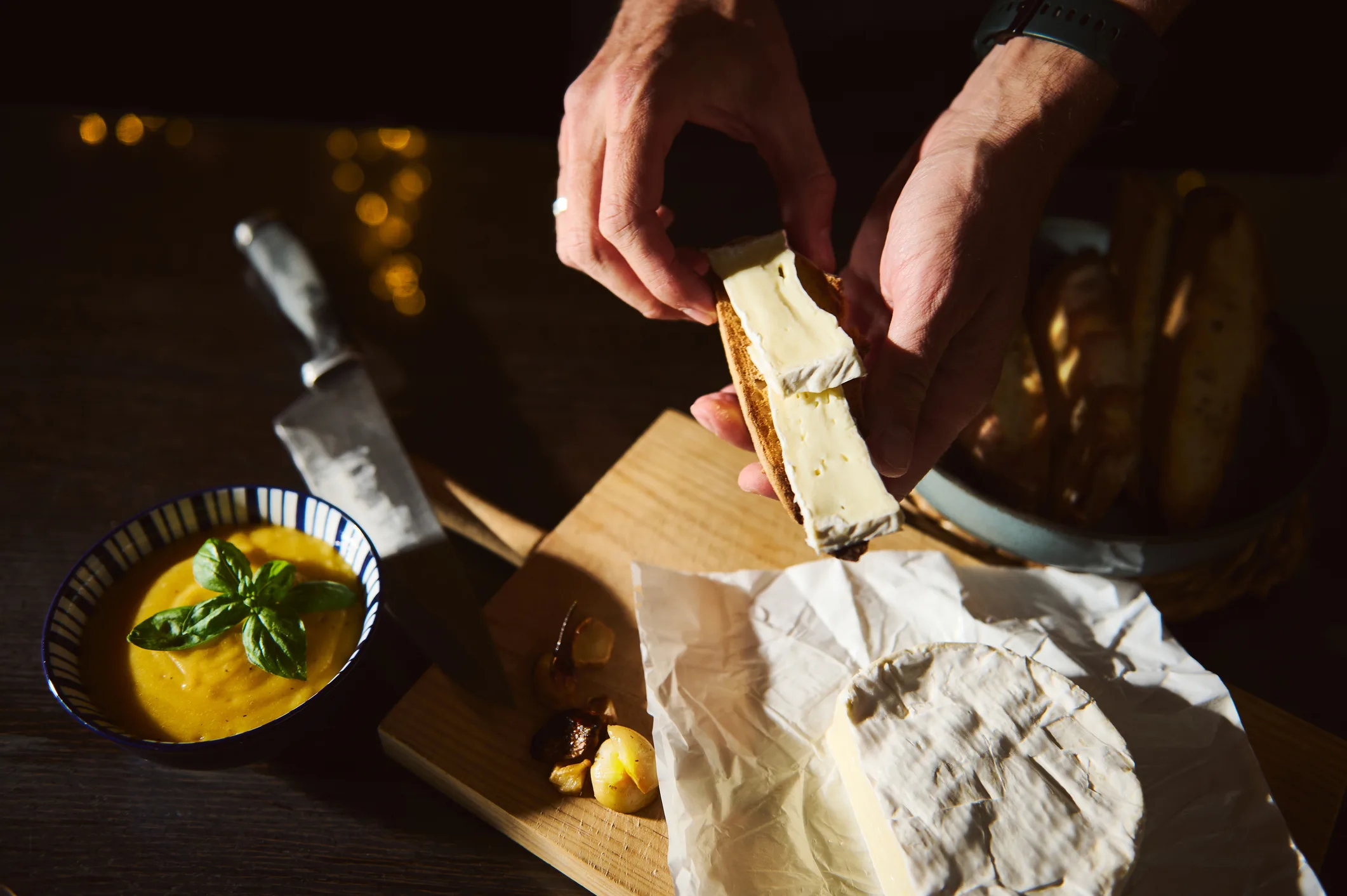 Hands Buttering Bread With Cream Cheese On a Wooden Board Beside Soup and Bread Basket