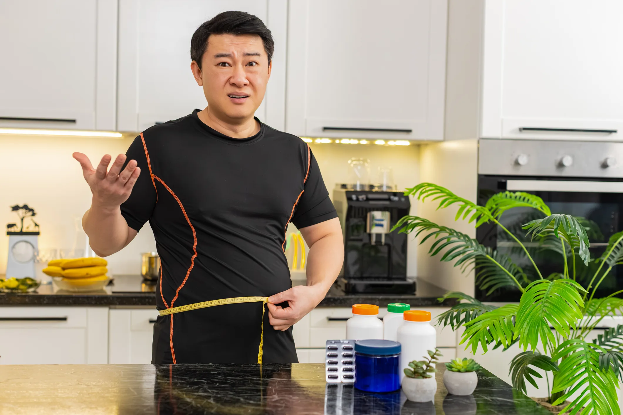 Heavy Asian adult man measures waist with tape upset about big belly fat weight gain at home kitchen