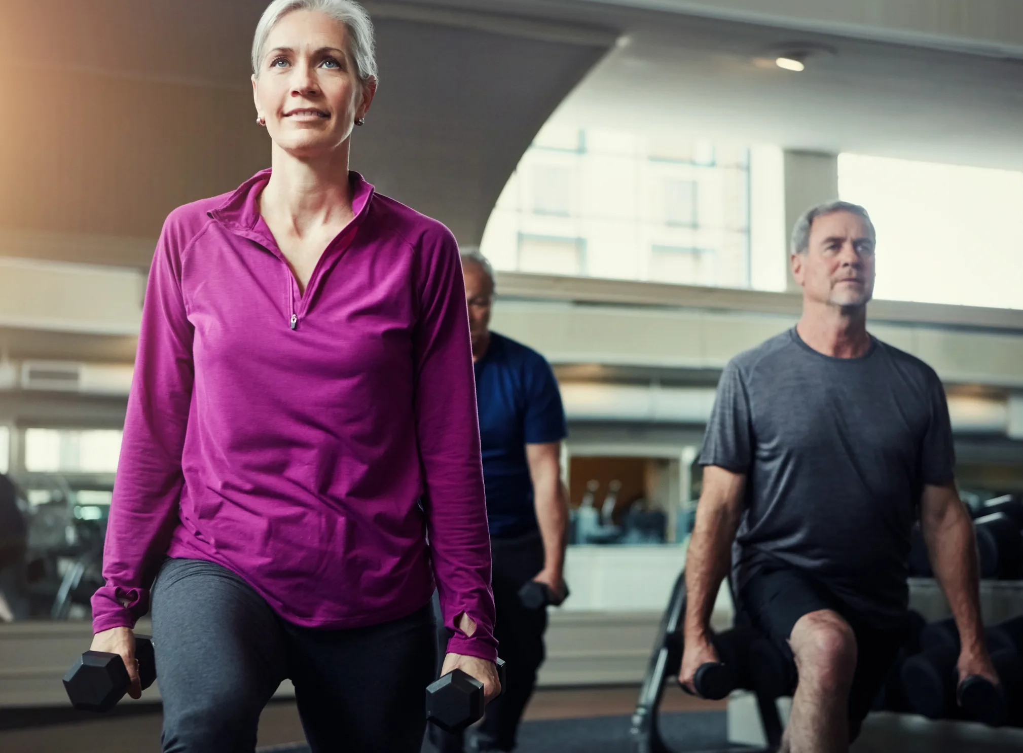 Woman, group and lunge with dumbbell in gym for fitness, coach or muscle development in class. Mature people, men and exercise for legs, health and balance for weightlifting workout at wellness club