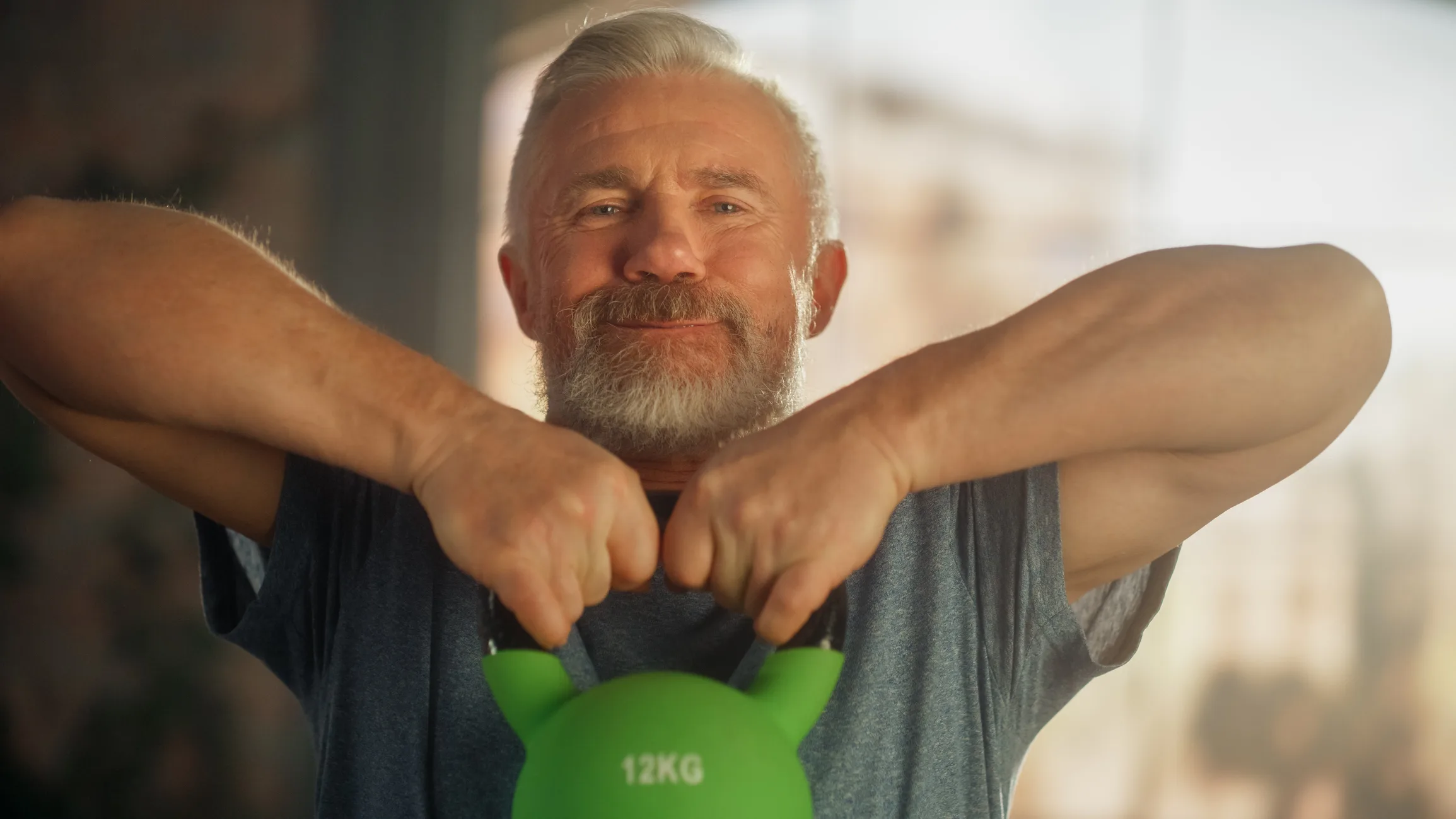 Portrait of a Happy Smiling Middle Aged Man Lifting a Heavy Kettlebell, Doing Core Strengthening Exercises During Morning Workout at Home in Sunny Apartment. Concept of Wellness and Fitness.