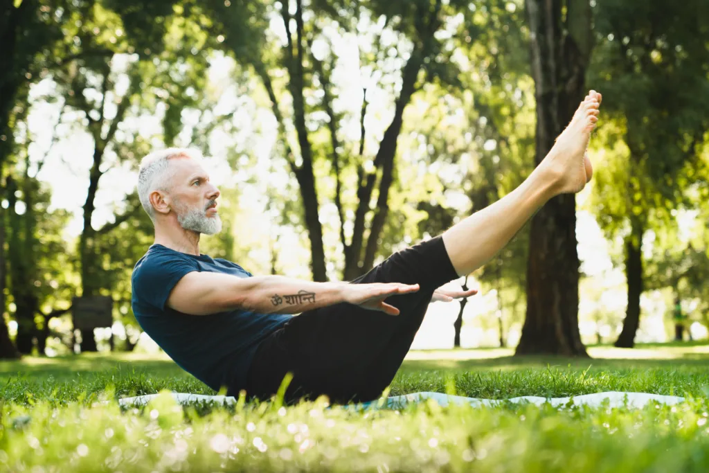Mature handsome man in sporty clothes doing workout practicing yoga fitness outdoors outside in park. Stretching exercises