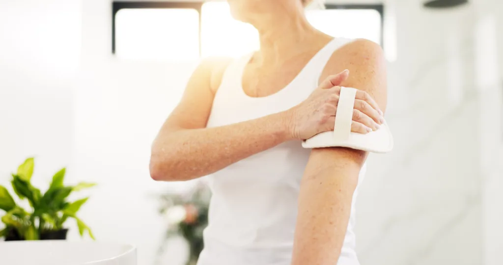 Home, skincare and old woman in bathroom, dry brush and lens flare with hygiene. Morning routine, pensioner and senior person with exfoliation, cleaning and wellness with healthy skin and body scrub