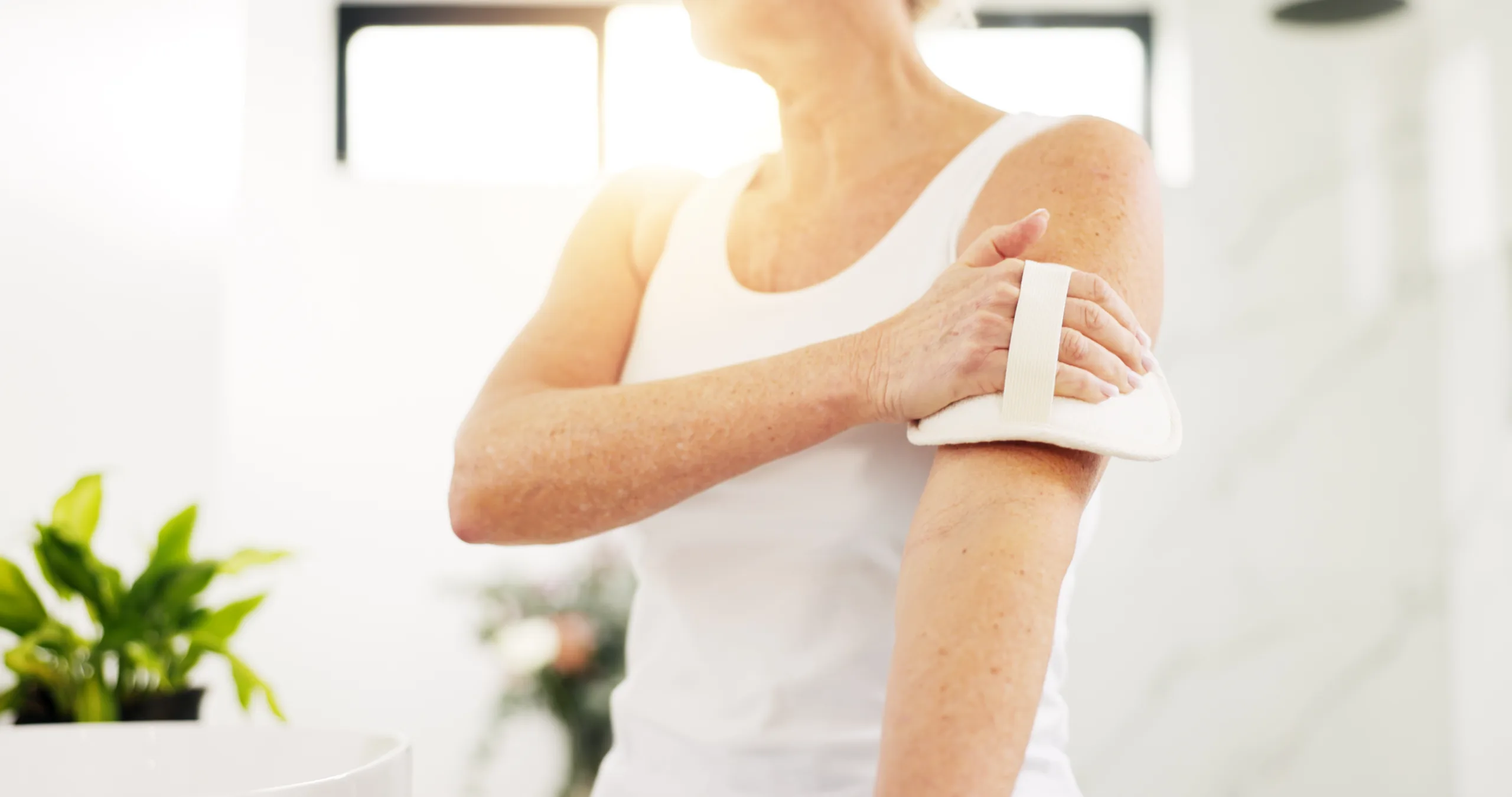 Home, skincare and old woman in bathroom, dry brush and lens flare with hygiene. Morning routine, pensioner and senior person with exfoliation, cleaning and wellness with healthy skin and body scrub