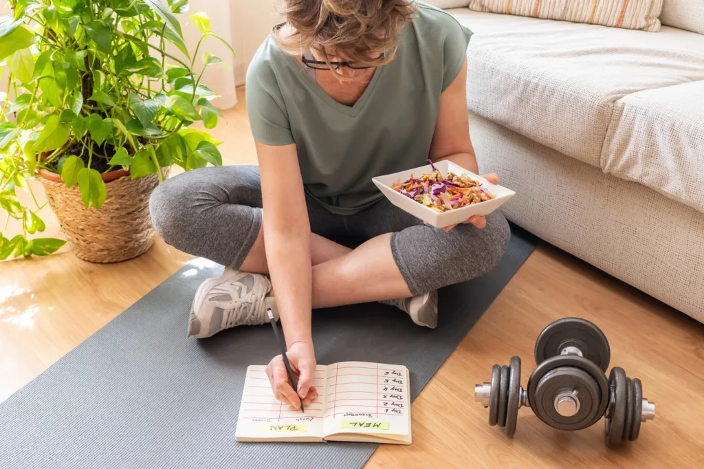 Mature woman sitting on a yoga mat is holding a healthy salad and writing down her meal and workout plan in a notebook, with dumbbells nearby