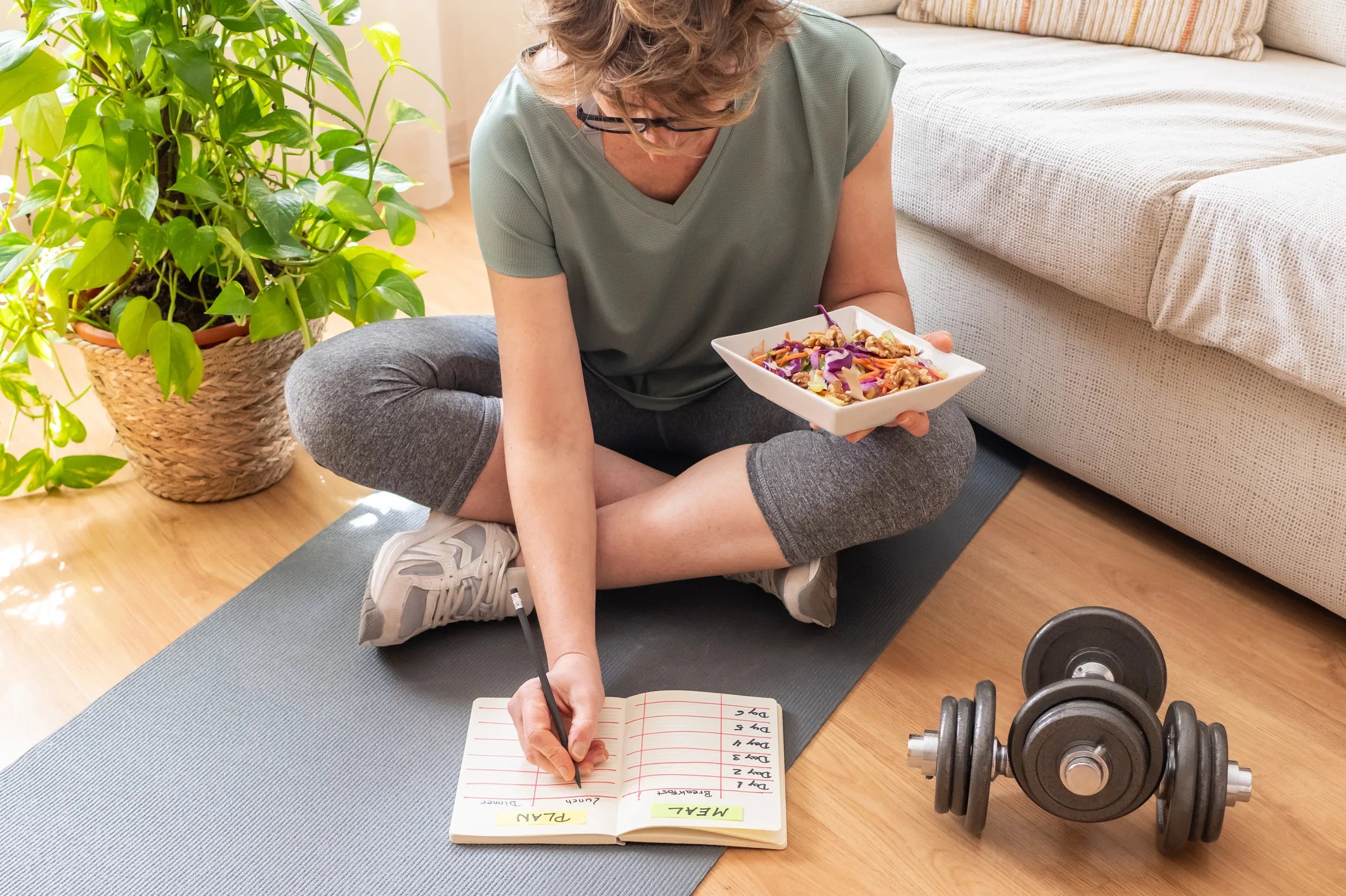 Mature woman sitting on a yoga mat is holding a healthy salad and writing down her meal and workout plan in a notebook, with dumbbells nearby