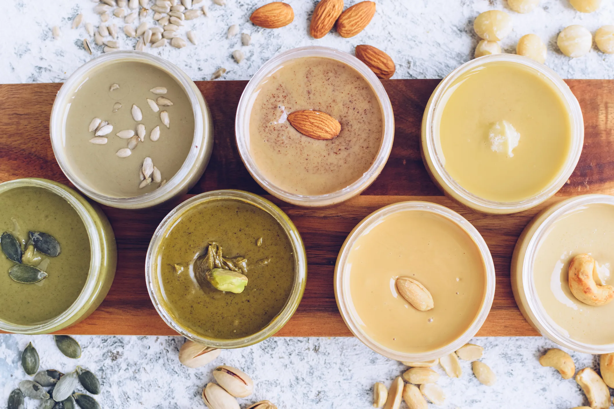Nuts and seeds butter in jars with ingredients. Homemade raw organic peanut, almond, hazlenut, cashew, pistachio, macadamia nuts paste and sunflower and pumpkin seeds butter on a table. Top view.