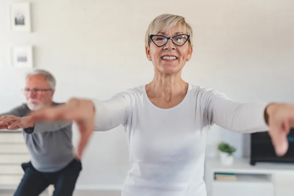 Smiling senior woman exercising at home, performing a squat with arms extended. In the background, an elderly man follows the workout in a bright and modern living room