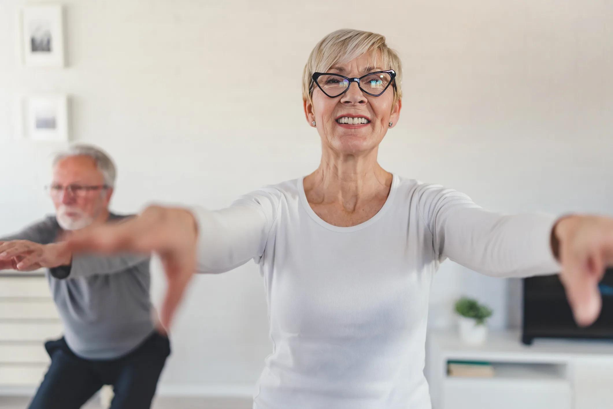 Smiling senior woman exercising at home, performing a squat with arms extended. In the background, an elderly man follows the workout in a bright and modern living room