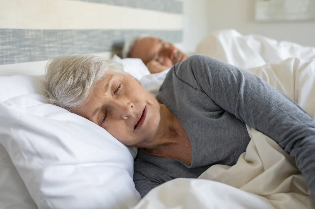 Old woman sleeping on bed at home with her husband. Elder lady sleeping in the bedroom with husband in background. Senior woman with grey hair wearing nightwear asleep in bed.