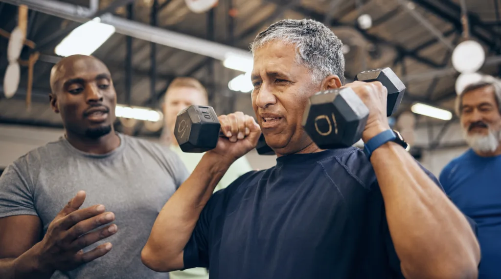 Fitness, old man with dumbbell and help from coach or personal trainer with weight lifting, strength and gym in retirement. Health, exercise and workout at senior sports club and coaching grandpa.