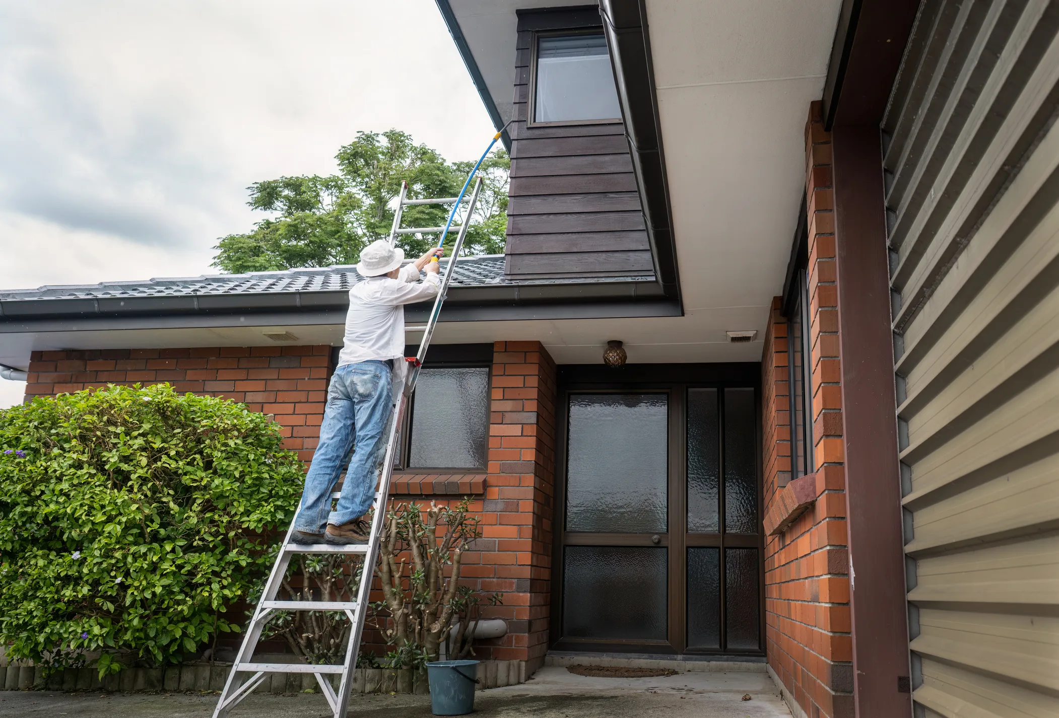 Man standing on the ladder and cleaning house with a long brush. Home maintenance work. Auckland.