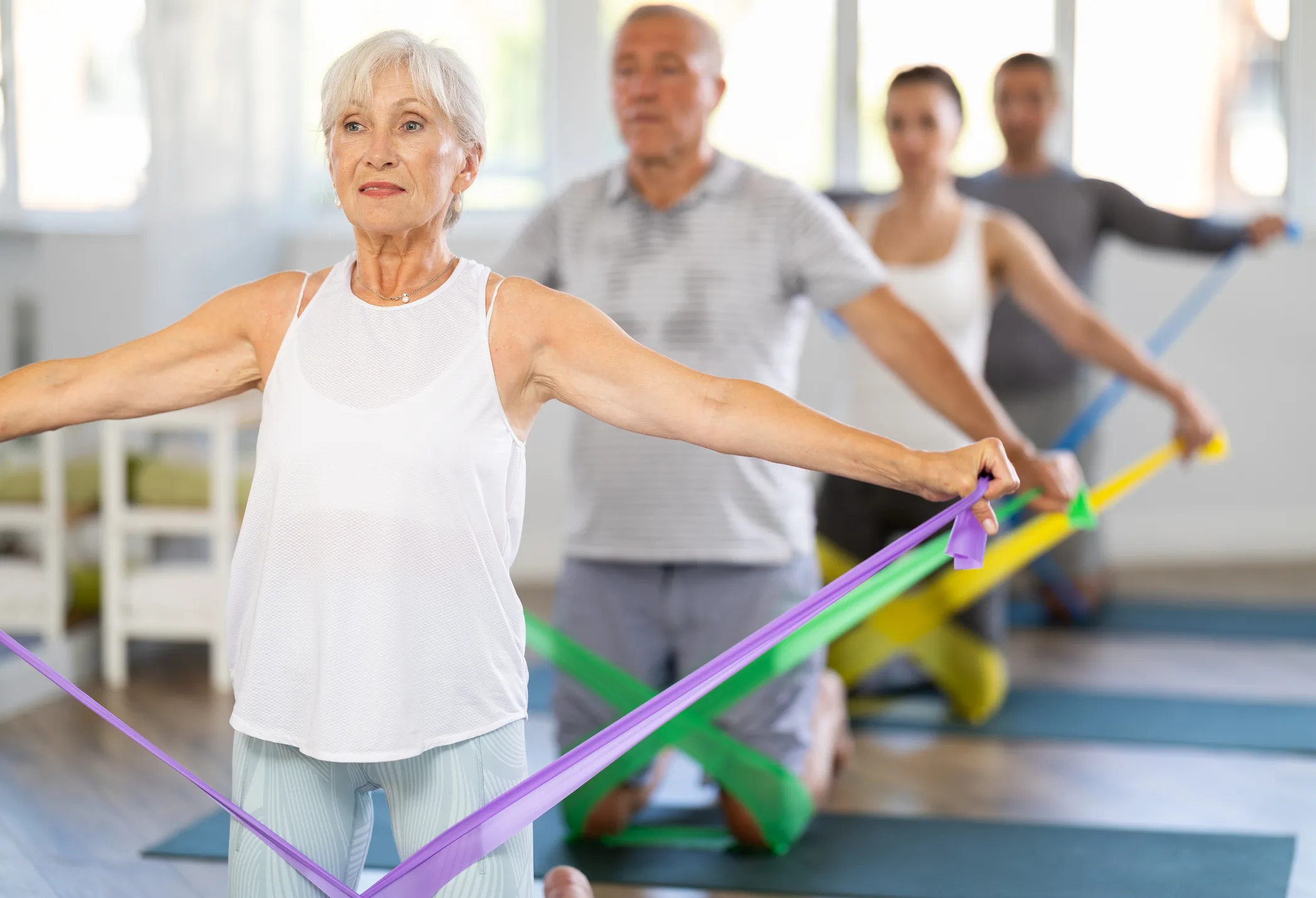 During training, mature woman kneeling performs exercises with colored rubber elastic band. She pulls on and releases expander, elastic band, do tasks to strengthen muscles of arms, shoulders, back