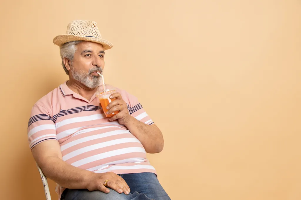 Retired senior indian man casual cloths and hat drinking soda water in plastic glass while sitting on chair relaxing isolated on beige studio background. Hot summer season, Retirement life.