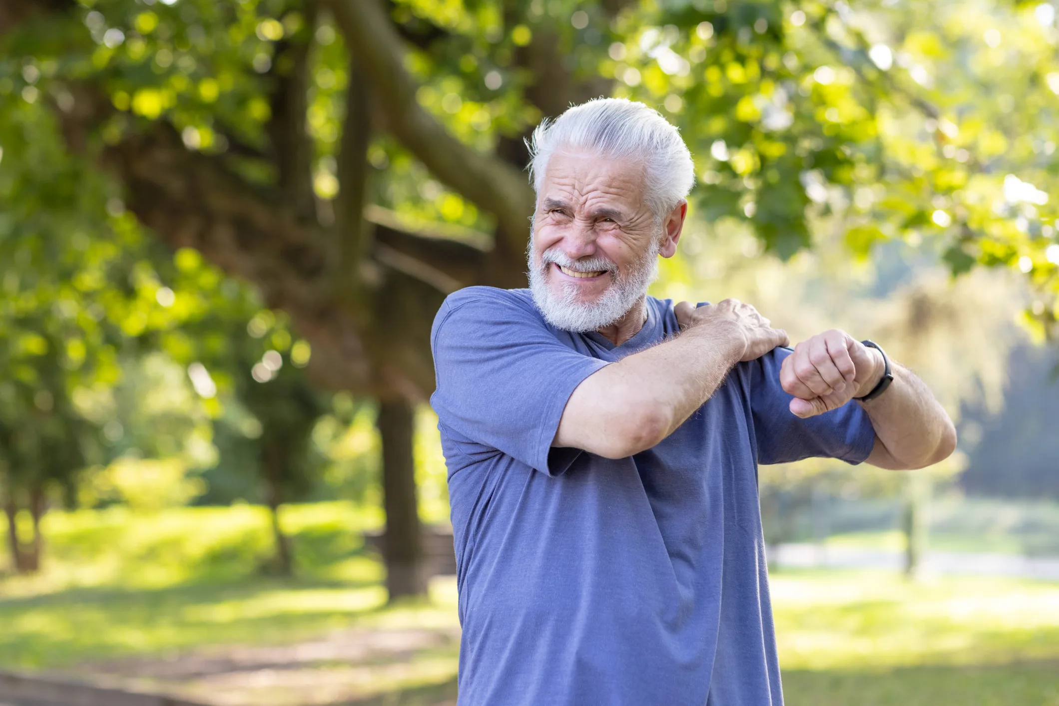 Senior gray-haired man sprained his ankle while walking in the park and exercising, standing outside and massaging his shoulder with his hand and feeling severe pain