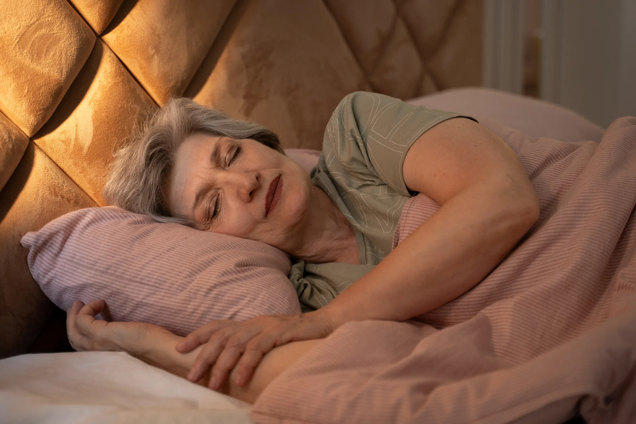 At night, an elderly woman can be seen sleeping soundly in bed. She is covered with a blanket, and her arms are wrapped around a pillow. The bed is made with pink sheets and a pink blanket.