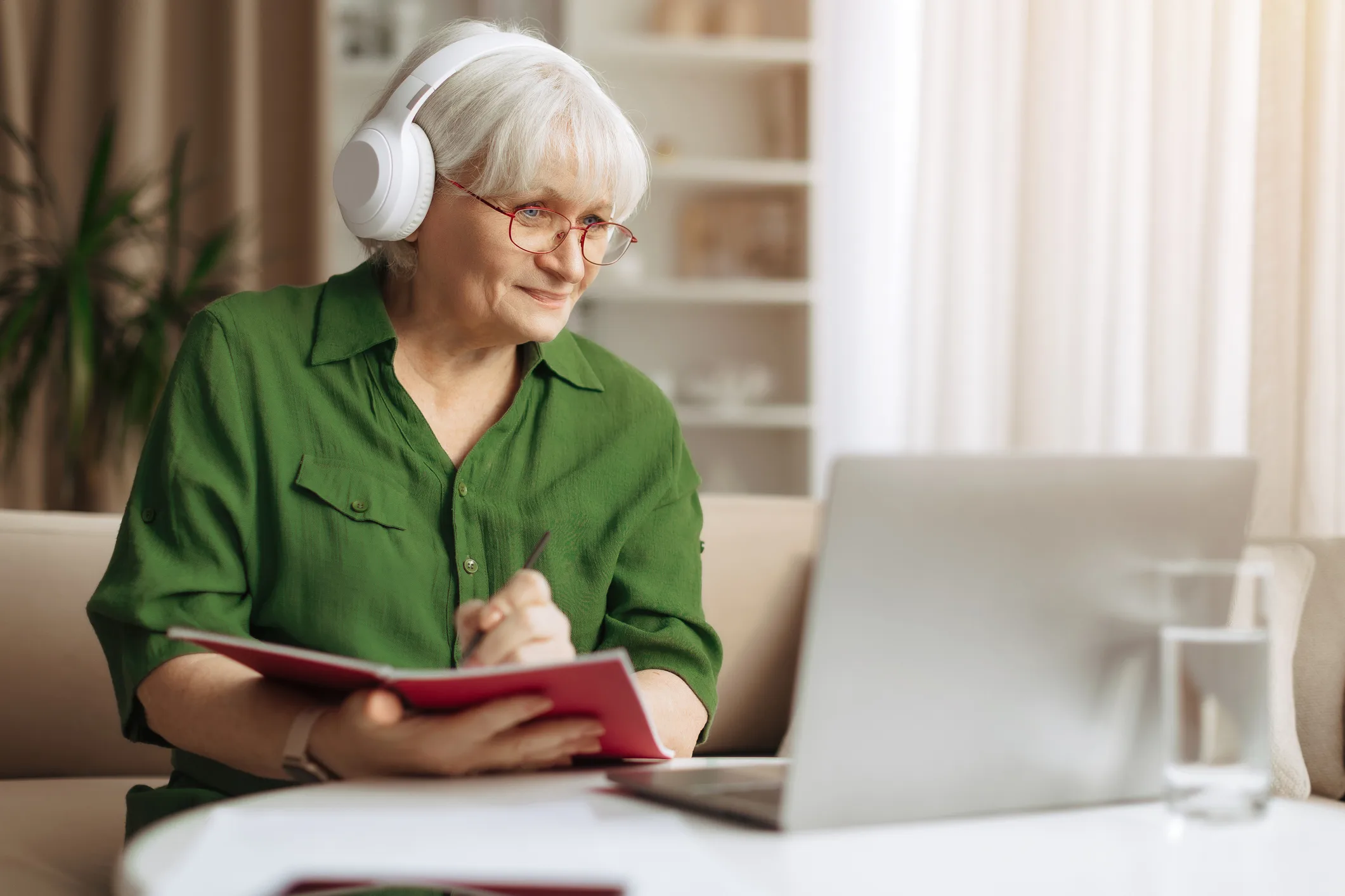 Elderly woman taking notes while wearing headphones and using laptop at home