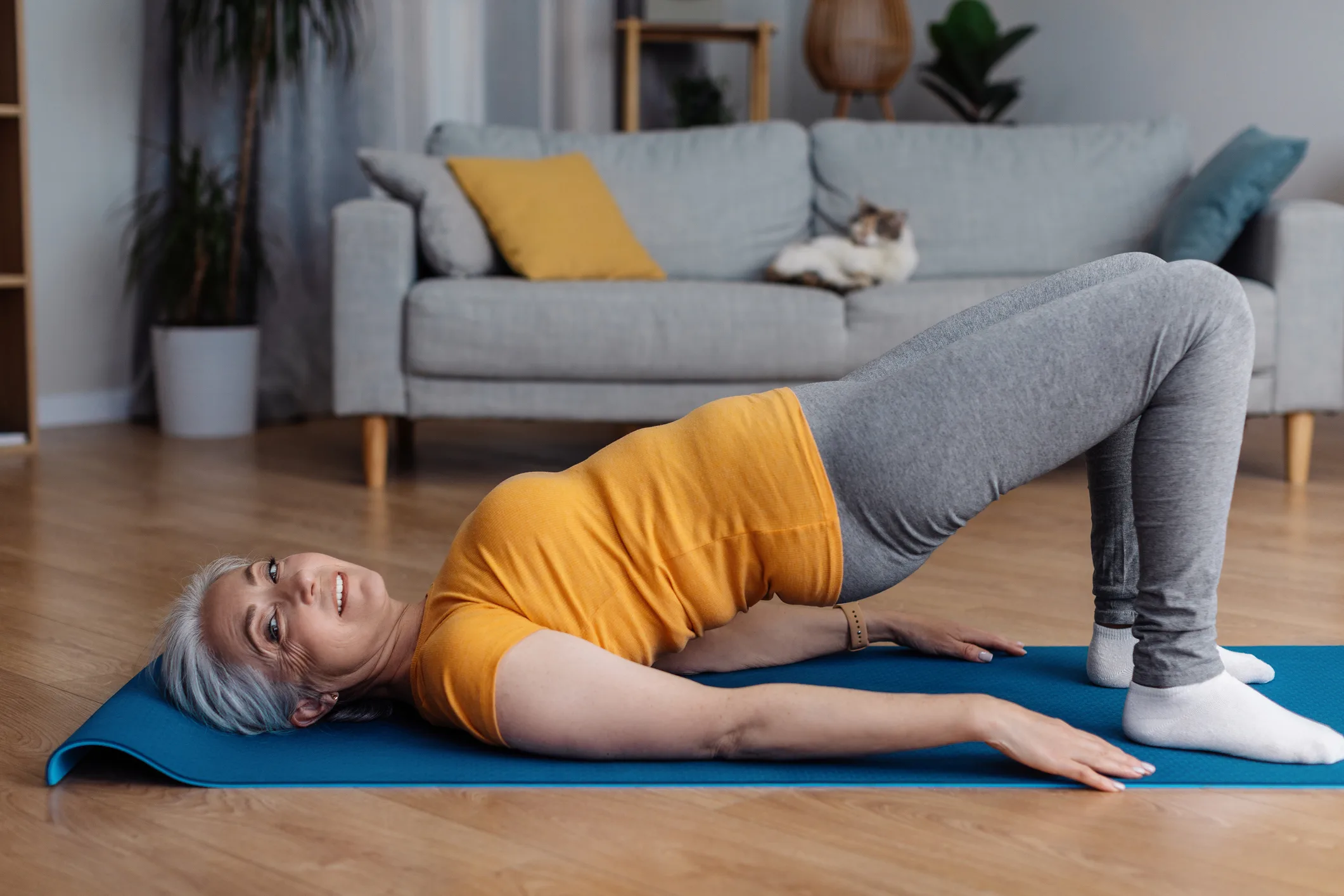 Sporty aged woman doing half bridge yoga pose, strengthening her abs muscles, training in living room, copy space