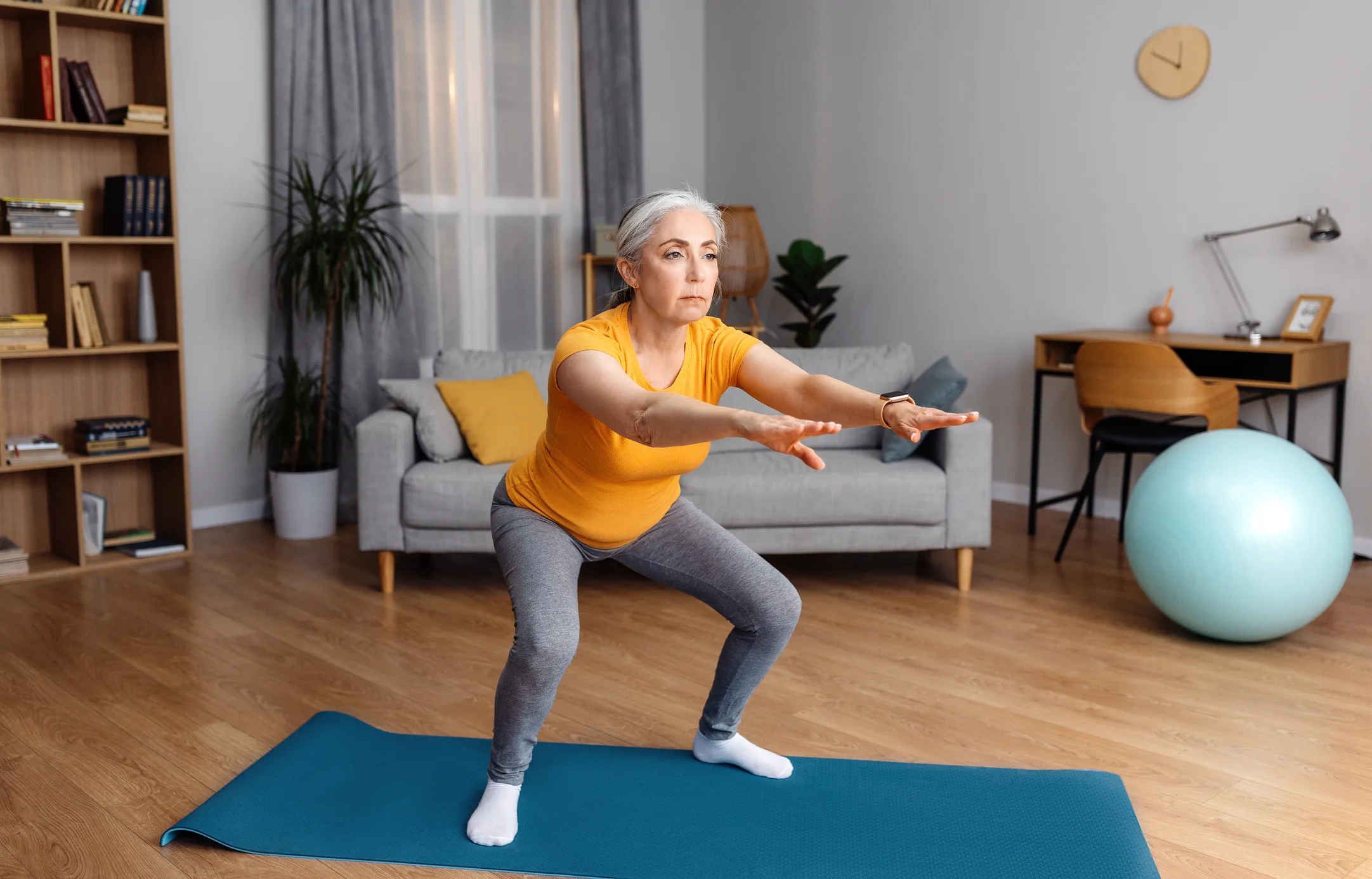 Home workout. Senior lady doing squats on yoga mat, working out at home in living room interior, free space. Fit aged woman exercising her legs on domestic training