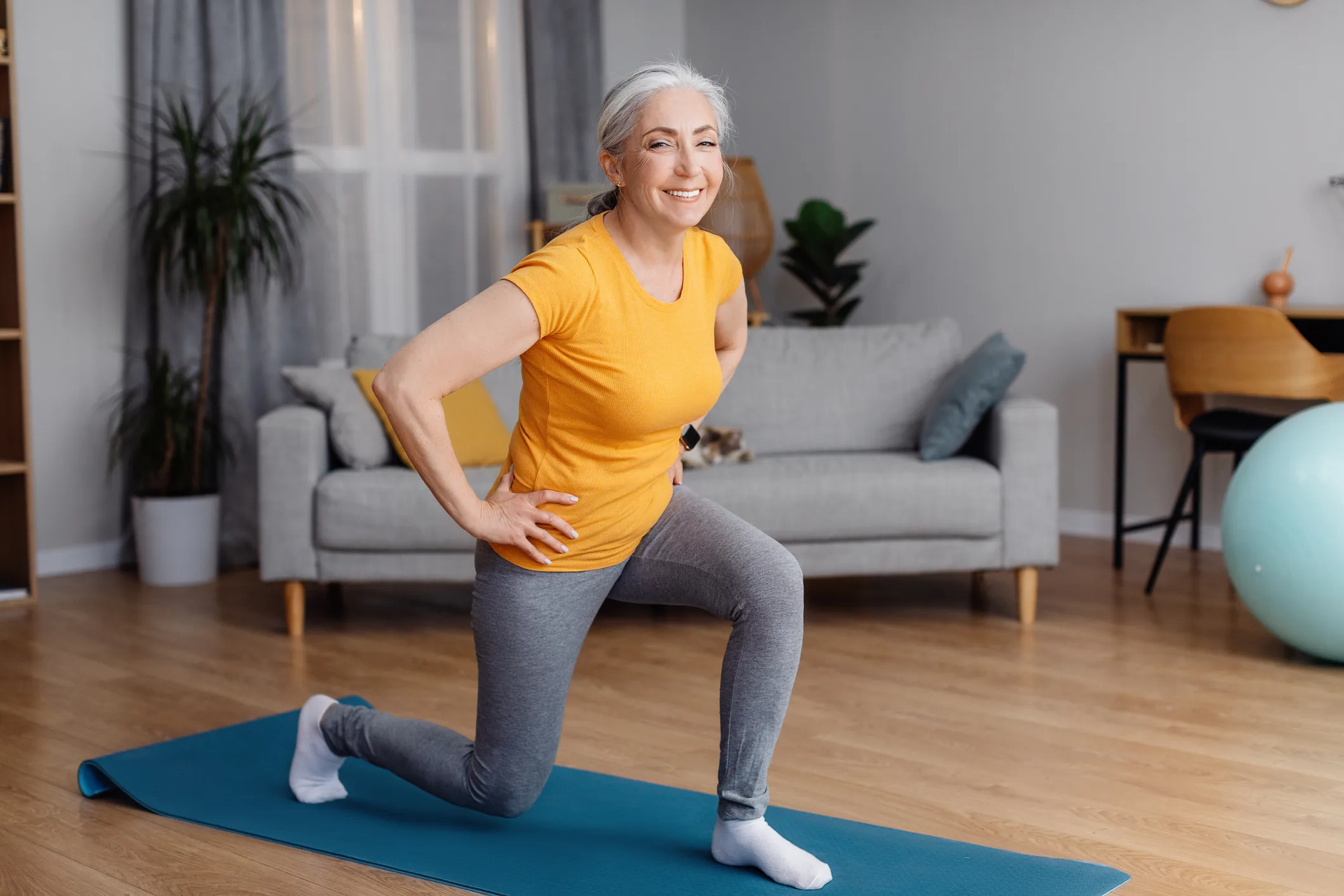Full length portrait of sporty senior woman keeping fit, exercising on yoga mat in living room and smiling at camera. Staying healthy on retirement