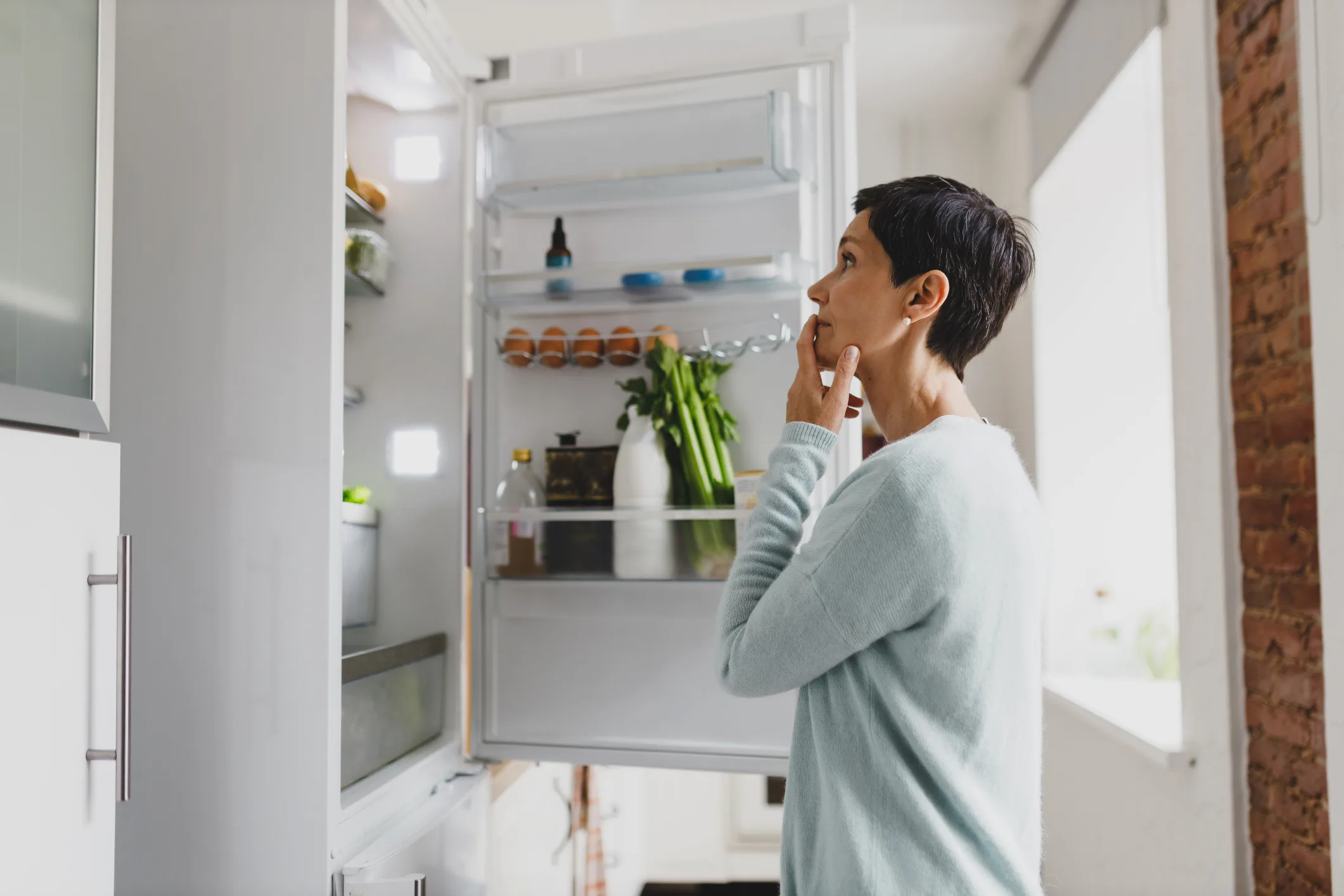 Side view of mature female with short hair standing in front of opened refrigerator at home with puzzled pensive facial expression, thinking of cooking breakfast, looking for ingredients